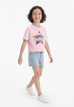 Smilende barn med skulderlangt krøllete hår har på seg en rosa "Sunset Heat" T-skjorte, lyse denimshorts og hvite joggesko mens det går mot hvit bakgrunn.