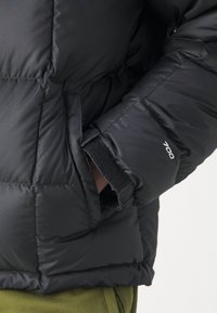 Person som bär en svart vadderad jacka med handen i sidofickan, ärmarna visar en etikett för "700" isolering, mot en enfärgad bakgrund.