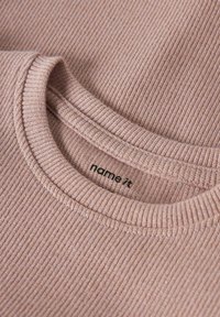 Camiseta de punto acanalado de color rosa claro con cuello redondo y etiqueta que muestra "name-it". La tela tiene una textura sutil y un brillo tenue.