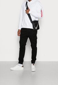 Persoon in witte hoodie, zwarte cargobroek, witte sneakers en een zwarte schoudertas met een groot wit Nike-logo.