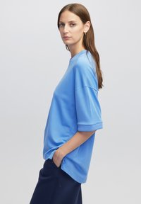 Světle modrý top s krátkým rukávem vyrobený z měkké tkaniny s volným střihem a ohrnutými manžetami, s kulatým výstřihem a bočními kapsami.