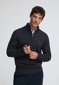 Dunkelgrauer Pullover mit Reißverschluss am Kragen, gerippten Bündchen und Saum. Verfügt über ein rotes Logodetail auf der Brust. Hergestellt aus einem weichen, strukturierten Stoff.