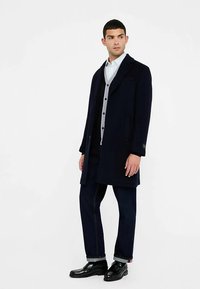 Tmavomodrý vlnený kabát s golierom, nosený cez sivú košeľu so zapínaním na gombíky, v kombinácii s tmavými džínsami a čiernymi leštenými topánkami.