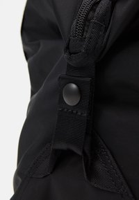 Gros plan sur un sac à dos en nylon noir, mettant en avant un tissu texturé, une fermeture éclair et un détail à bouton-pression, avec une couture renforcée sur les bretelles.