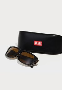 Occhiali da sole rettangolari con lenti marroni e montatura nera e tartaruga accanto a un astuccio nero con il logo Diesel rosso e bianco.