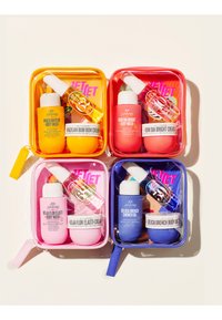 Fire farverige rejse-størrelse selvplejesæt med body wash, cremer og sprays, hver i klare lynlåsposer i gul, rød, pink og blå.