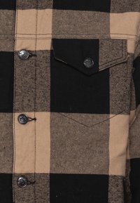 Rutet flannel skjorte i sort og tan. Inkluderer trykknapper, brystlomme og struktureret stof. Designet har store vekslende tern.
