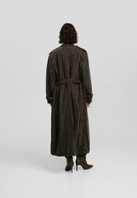 Długi brązowy płaszcz trench z paskiem w talii, z guzikami przy mankietach i lekko wyblakłą fakturą. Projekt zawiera naramienniki.