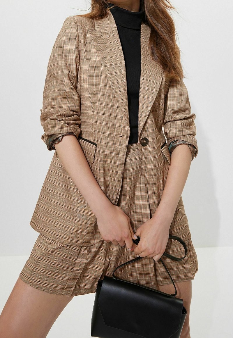 Femme portant un blazer à carreaux beige et un short assorti avec un col roulé noir, tenant un petit sac à main noir par sa sangle.