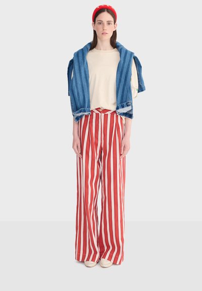 Femme portant un pantalon à jambes larges à rayures verticales rouges et blanches, une chemise crème, une veste en jean posée sur les épaules, un bandeau rouge et des chaussures blanches.
