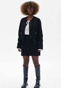 Kobieta z kręconymi włosami, ubrana w czarną zapinaną kurtkę i spódnicę, białą bluzkę, duże okulary przeciwsłoneczne oraz czarne botki do kolan, stojąca na tle białym.