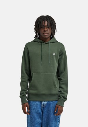 Giovane uomo dalla pelle scura e con dreadlocks, che indossa una felpa verde con cappuccio e jeans blu, in piedi contro uno sfondo chiaro e semplice.