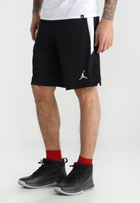 Schwarze Sportshorts mit einem weißen Seitenpanel, versehen mit einem weißen Jumpman-Logo. Getragen mit schwarzen High-Top-Sneakern und roten Socken.