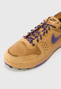 Hnedé tenisky s purpurovými akcentmi, zsuede a sieťovaného materiálu, okrúhla špička, štruktúrovaný povrch a purpurové šnúrky. Logo Nike na boku.