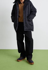 Zwarte gewatteerde parka met capuchon, oranjebruine polo met kraag, losse zwarte broek en marineblauwe schoenen met bruine zolen op een groene achtergrond.