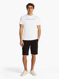 Hvid bomulds t-shirt med "TOMMYJEANS" logo, parret med sorte cargo shorts og cremede sneakers. Model står med hænderne i lommen.
