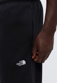 Gros plan sur la main d'une personne reposant à côté d'un pantalon de survêtement noir avec un logo blanc The North Face sur la cuisse.