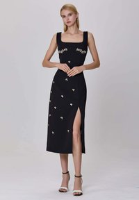 Jeune femme dans une robe midi noire avec des embellissements floraux et une fente haute, portant des sandales à talons hauts blanches et noires sur un fond uni.