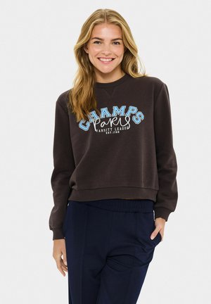 Brauner Pullover mit dem Schriftzug "CHAMPS PARIS" in Blau und Weiß, Rundhalsausschnitt, lockere Passform und gerippte Bündchen. Kombiniert mit marineblauen Hosen.