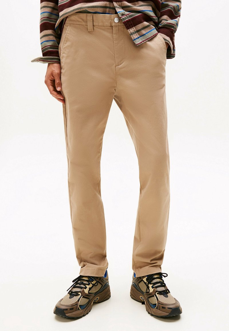 Homme portant un pantalon beige, une chemise à manches longues rayée aux tons marron et bleu, et des chaussures de sport marron avec des lacets noirs.