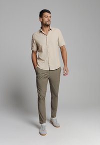 Beige overhemd met korte mouwen en knopen, gemaakt van een gladde stof, gecombineerd met olijfgroene slim-fit broek en witte sneakers met rubberen zolen.
