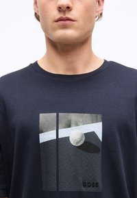 Άνδρας που φοράει μπλε σκούρο πουκάμισο με γραφικό μια λευκή μπάλα του τένις σε γήπεδο και τη λέξη "BOSS" με μαύρα γράμματα.