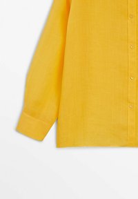 Chemise jaune à manches longues avec poignets visibles et partie de la patte de boutonnage sur fond blanc uni.