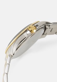 Montre en acier inoxydable avec une lunette et une couronne rehaussées d'or. Fini lisse et brillant avec un bracelet métallique ajustable et un cadran circulaire.