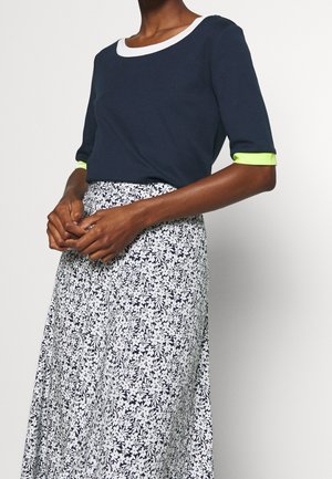 Henkilö käyttää tummansinistä yläosaa, jossa on valkoinen kaulus ja neonvihreät hihansuut, yhdistettynä korkeavyötäröiseen valkoiseen hameeseen, jossa on musta kukkakuosi.