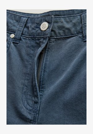 Tamsiai mėlyni džinsiniai kelnės su dalinai atidarytu užtrauktuku ir sidabriniu mygtuku, pažymėtu užrašu „NEXT COLLECTION“.