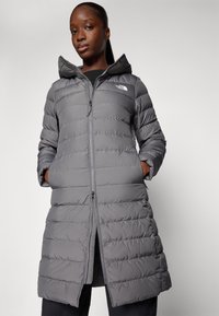 Pelēks pūkains mētelis ar vertikālām šuvēm, regulējamu kapuci, rāvējslēdzēja aizdari un sānu kabatām. Uz krūtīm ir The North Face logo.