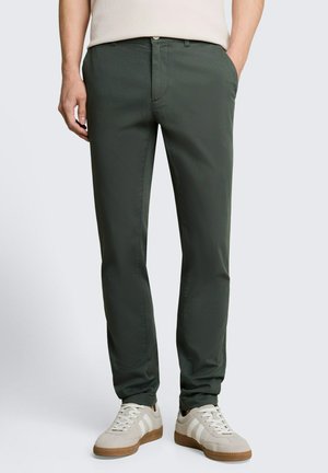 Mann trägt dunkelgrüne Slim-Fit-Hose mit weißen Sneakers und einem hellbeigen ärmellosen Oberteil, Hände in den Taschen.