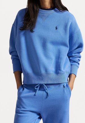 Femme portant un sweat-shirt bleu et un pantalon de survêtement assorti avec un cordon de serrage et un petit logo brodé sur la poitrine et la poche. Mains dans les poches.
