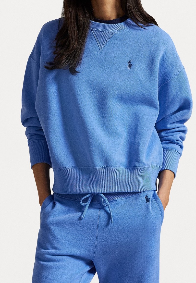 Femme portant un sweat-shirt bleu et un pantalon de survêtement assorti avec un cordon de serrage et un petit logo brodé sur la poitrine et la poche. Mains dans les poches.