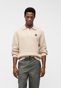 Νέος άνδρας που φοράει μπεζ ριγωτό πουλόβερ με γιακά και σκούρο γκρι παντελόνι με κορδόνι, στέκεται μπροστά σε απλό λευκό φόντο.