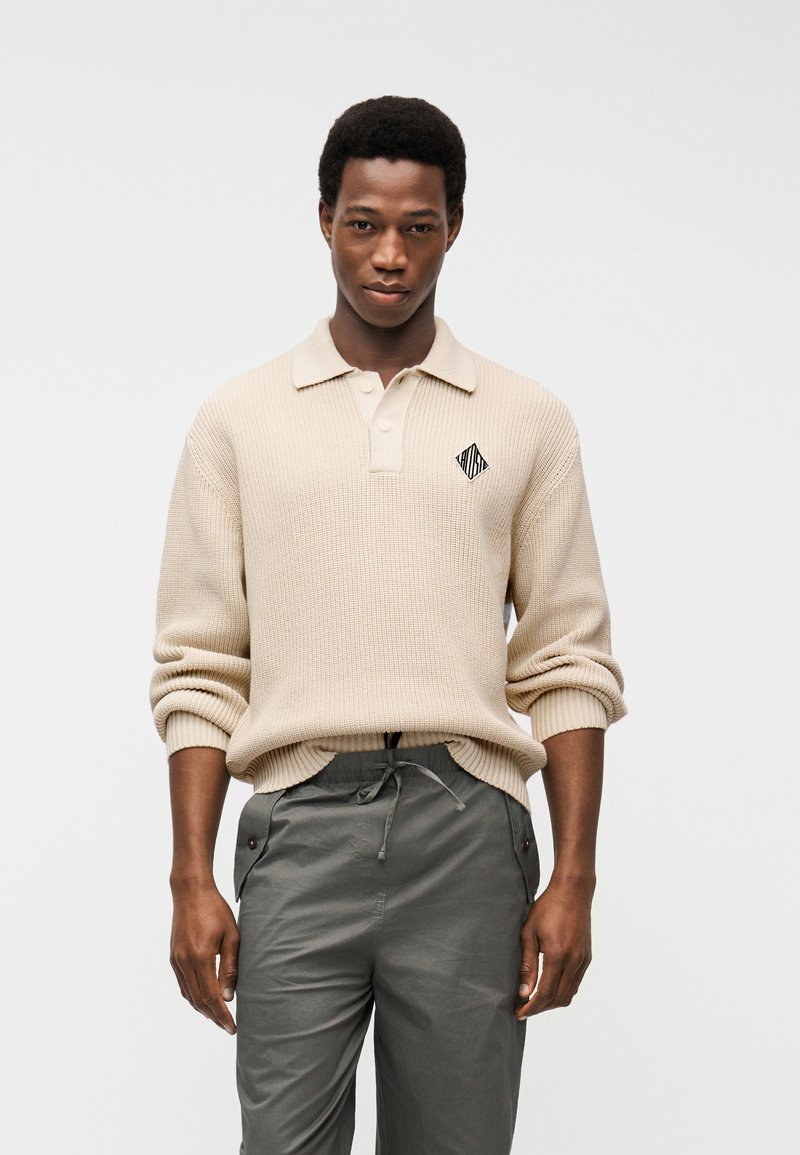 Νέος άνδρας που φοράει μπεζ ριγωτό πουλόβερ με γιακά και σκούρο γκρι παντελόνι με κορδόνι, στέκεται μπροστά σε απλό λευκό φόντο.