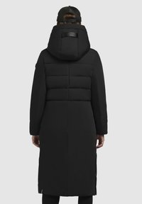 Crna puffer jakna dugog kroja, s horizontalnim šavovima na leđima, velikim kapuljačom i podešavanjem na izrezu.