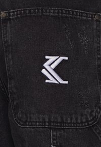 Czarny denims z lekko postrzępioną teksturą, z wyraźnym białym wyszywanym logo na tylnej kieszeni.