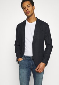 Blazer azul-marinho com um único botão, lapelas recortadas e dois bolsos frontais, usado sobre uma t-shirt branca e combinado com jeans azuis desgastados.