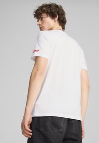 Hvid bomulds T-shirt med rund halsudskæring og korte ærmer. Har et rødt logo på venstre ærme. Stylet med mørke bukser.