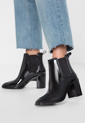 Bottines noires brillantes à talons blocs portées avec un jean bleu clair court à ourlet effiloché sur fond blanc.