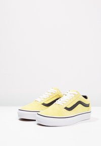 Paire de baskets basses en daim jaune avec lacets blancs, bande latérale noire et semelles épaisses en caoutchouc blanc sur une surface blanche.