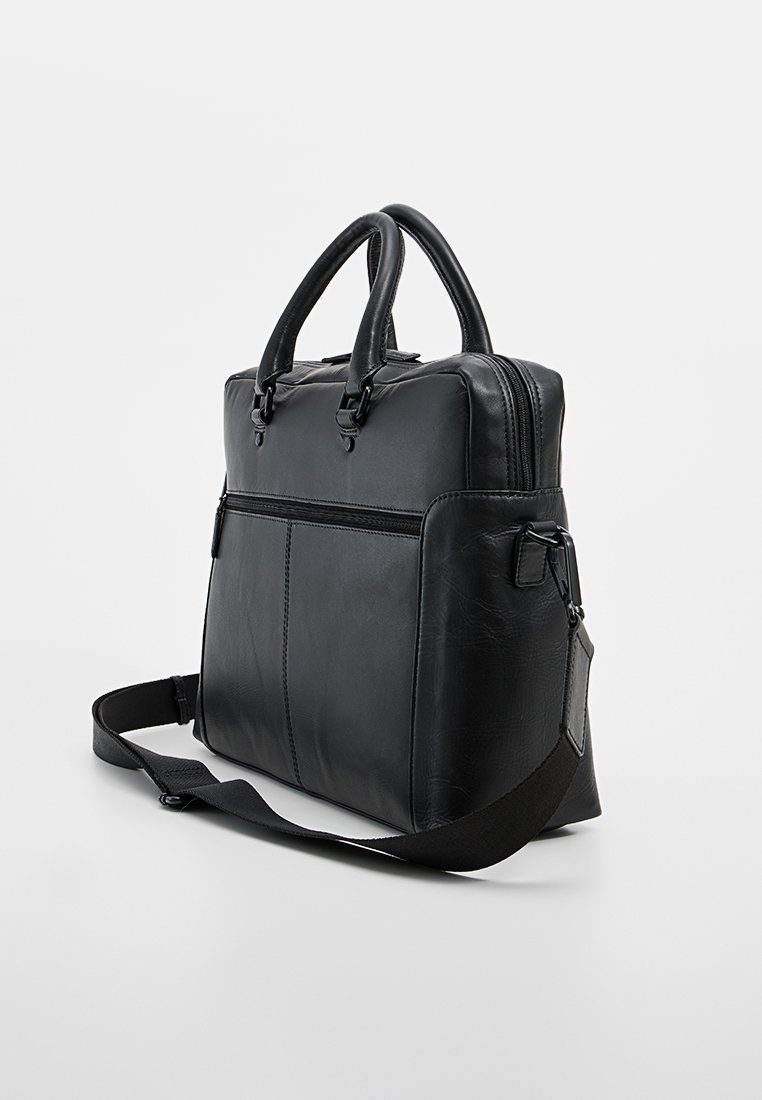Sac en cuir noir avec deux poignées supérieures, une bandoulière amovible, une fermeture éclair et une poche avant. Texture lisse et design minimaliste.