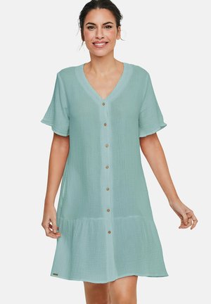Femme souriante, portant une robe chemise à manches courtes bleu clair avec un col en V et un ourlet à volants.