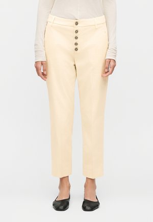 Γυναίκα που φοράει κρεμ παντελόνι με ψηλή μέση και πέντε διακοσμητικά κουμπιά στο μπροστινό μέρος, σε συνδυασμό με μαύρα επίπεδα παπούτσια και μπεζ τοπ με μακριά μανίκια.