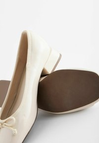 Chaussures en cuir verni beige avec un bout rond, un petit talon et un nœud décoratif. La semelle est marron avec une texture striée.