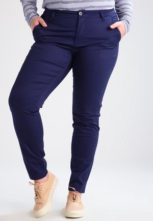 Persoon draagt marineblauwe slim-fit broek, lichtbeige sneakers en een grijs gestreept shirt met lange mouwen, met handen in de zakken.