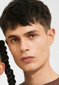 Modèle masculin avec des cheveux courts et foncés et une peau claire. Il porte une petite boucle d'oreille. Gros plan, mettant l'accent sur le visage et le haut du torse, fond neutre.