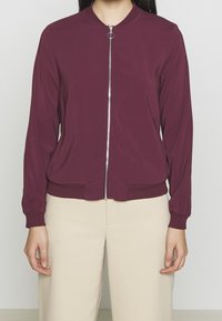 Burgundská bomber bunda na zip s žebrovanými manžetami a spodním lemem, s kulatým límcem a hladkým povrchem. Nositá přes světle béžové kalhoty.