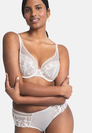 Mujer de piel morena clara vistiendo un sujetador de encaje blanco translúcido y ropa interior a juego, con los brazos cruzados sobre el torso, frente a un fondo liso.